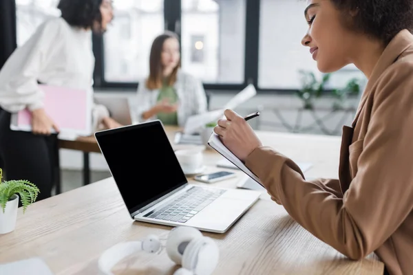 Jovem empresária afro-americana escrevendo em notebook perto de laptop com tela em branco no escritório — Fotografia de Stock