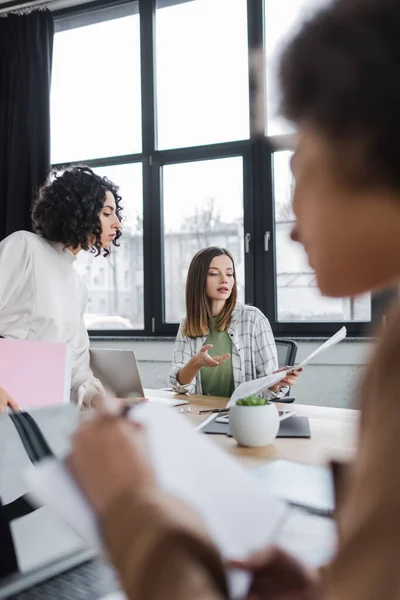 Businesswoman pointing at newspaper near laptop and muslim colleague in office — Photo de stock