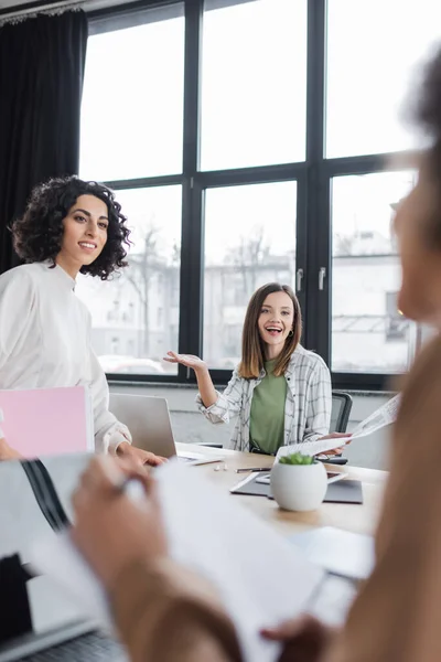 Positive multiethnic businesswoman holding papers near blurred african american colleague in office — Fotografia de Stock