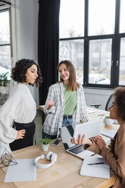 Fröhliche Geschäftsfrau im Gespräch mit multiethnischen Kollegen in der Nähe von Geräten im Büro — Stockfoto