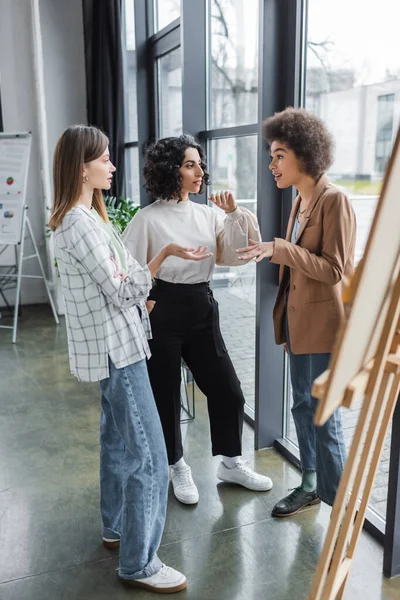 Multicultural businesswomen talking near blurred board in office — стоковое фото