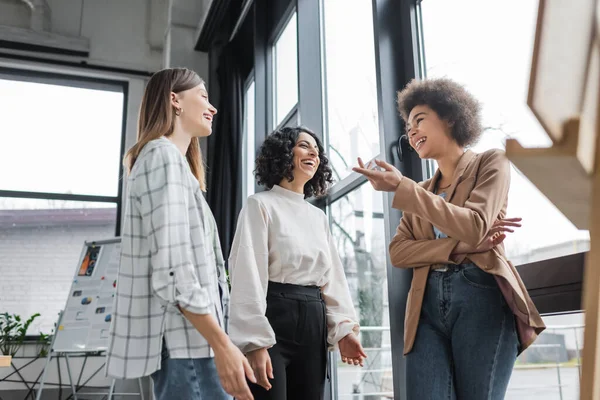 Vue en angle bas des femmes d'affaires interraciales heureuses riant au bureau — Photo de stock