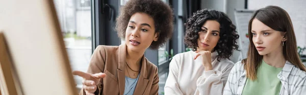 African american businesswoman pointing at blurred board near multiethnic colleagues in office, banner — Stockfoto