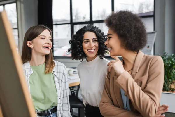 Cheerful interracial businesswoman talking near blurred board in office — Fotografia de Stock