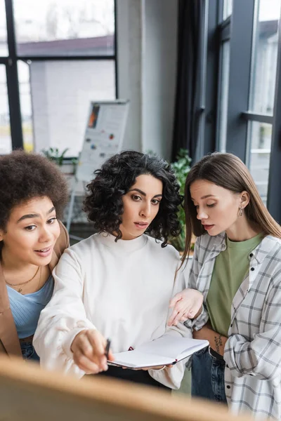 Muslim businesswoman holding notebook near interracial colleagues and blurred board in office - foto de stock