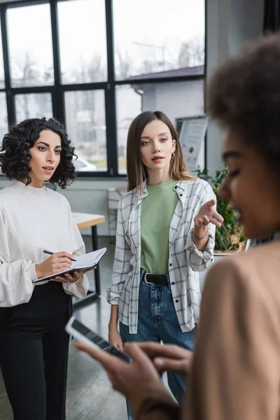 Empresarias interraciales con cuaderno hablando con un borroso colega afroamericano con celular en la oficina - foto de stock