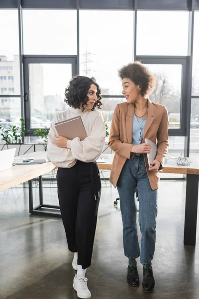 Positive interracial businesswomen with paper folders and laptop in office — Stockfoto