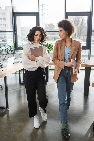 Femme d'affaires afro-américaine tenant un ordinateur portable et parlant à un collègue arabe avec des documents en fonction — Photo de stock