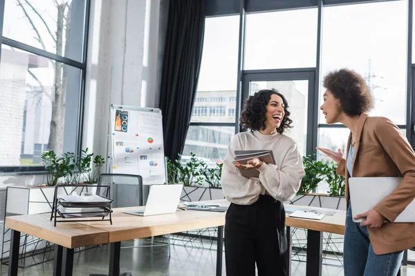 Heureuses femmes d'affaires multiethniques avec ordinateur portable et papiers parlant au bureau — Photo de stock