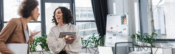 Mulheres de negócios multiétnicas alegres com papéis e laptop conversando no escritório, banner — Fotografia de Stock