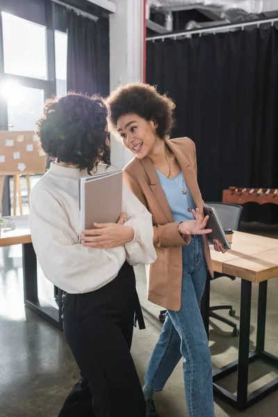 Donna d'affari afro-americana sorridente che tiene in mano un portatile e parla con un collega arabo con cartelle di carta in ufficio — Foto stock