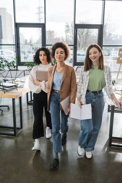 Happy interracial businesswomen with papers and laptop walking in office — стоковое фото
