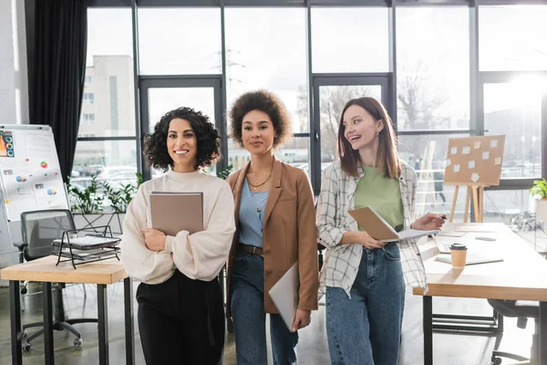 Cheerful multicultural businesswomen with papers, notebook and laptop in office — стоковое фото