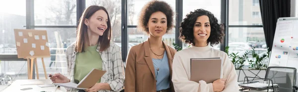 Positive multicultural businesswomen with notebook and documents in office, banner — Stock Photo