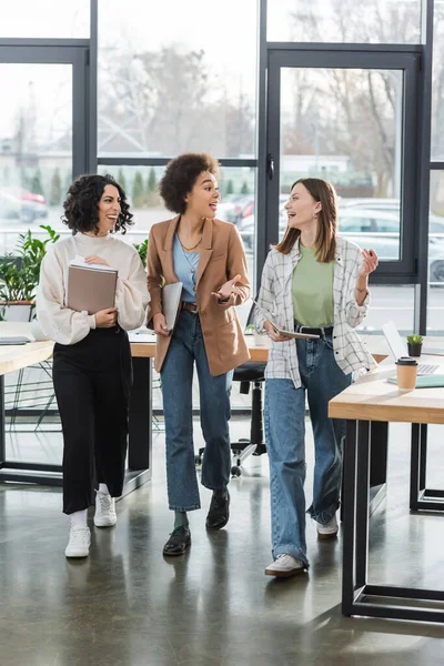 Positive multicultural businesswomen with laptop and documents talking while walking in office — Foto stock