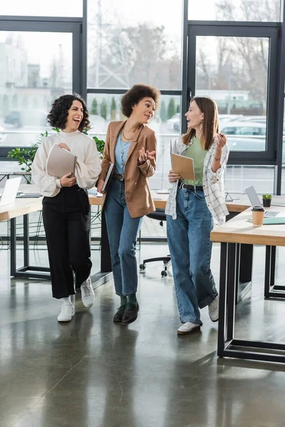 Cheerful multicultural businesswomen with documents walking in office — Stock Photo