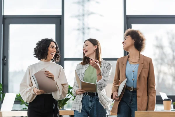 Cheerful multiethnic businesswomen with documents and laptop talking in office — Photo de stock