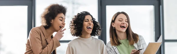 Laughing multiethnic businesswomen looking at notebook in office, banner — Foto stock