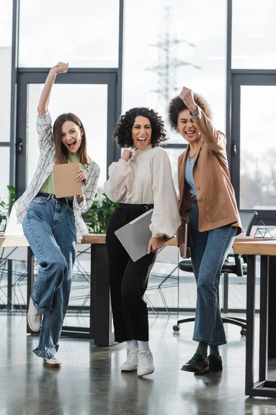 Excited multiethnic businesswomen with laptop and papers looking at camera in office - foto de stock