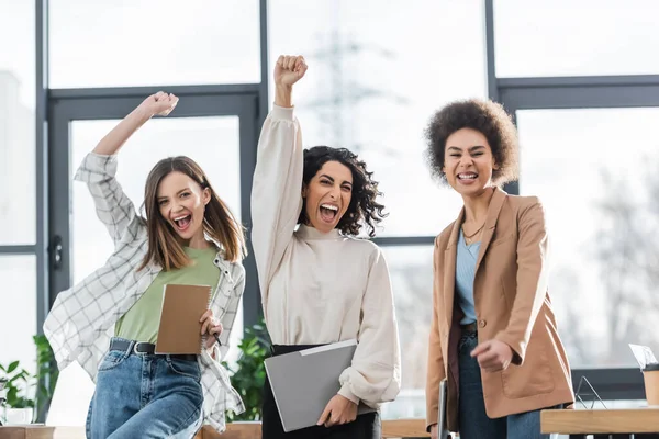 Excited multicultural businesswomen with papers and laptop showing yes gesture in office — Stock Photo