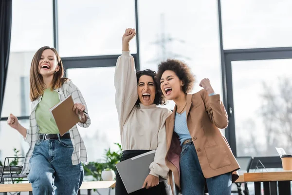 Aufgeregte multikulturelle Geschäftsfrauen mit Papieren, die Ja-Geste im Amt zeigen — Stockfoto