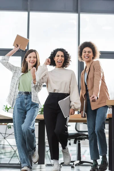 Excited multicultural businesswomen with papers and laptop showing yes gesture in office - foto de stock