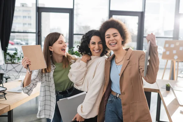 Positive interracial businesswomen with laptop and papers standing in office — Stock Photo