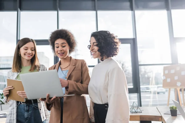 Empresarias multiétnicas positivas mirando el ordenador portátil en la oficina — Stock Photo
