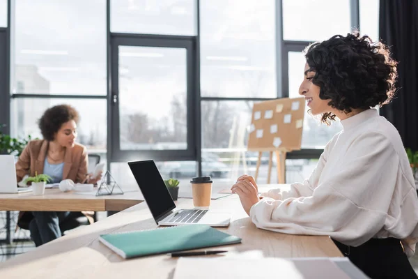 Sorridente donna d'affari musulmana che guarda il computer portatile vicino a carte e caffè per andare in ufficio — Foto stock