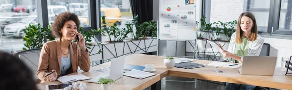 Positive african american businesswoman talking on cellphone near colleague in office, banner - foto de stock