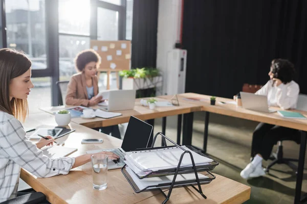 Junge Geschäftsfrau benutzt Laptop in der Nähe von Dokumenten und Wasser im Büro — Stockfoto