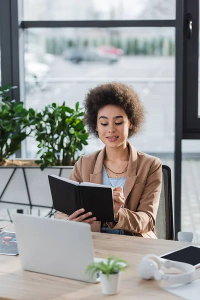 Empresaria afroamericana positiva sosteniendo portátil cerca de dispositivos y auriculares en la oficina - foto de stock