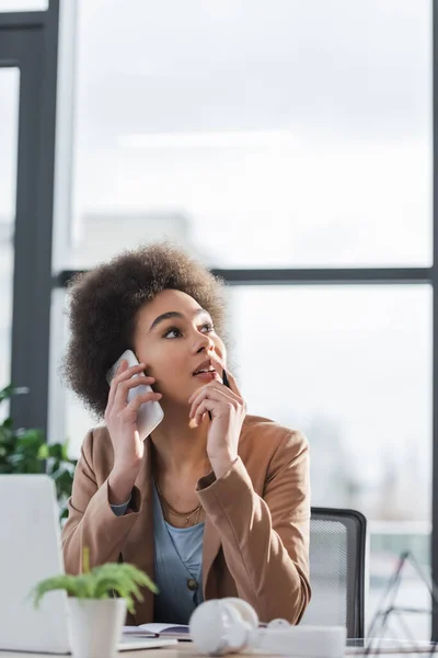 Pensativa mujer de negocios afroamericana hablando en el teléfono inteligente cerca de la computadora portátil y auriculares en la oficina - foto de stock