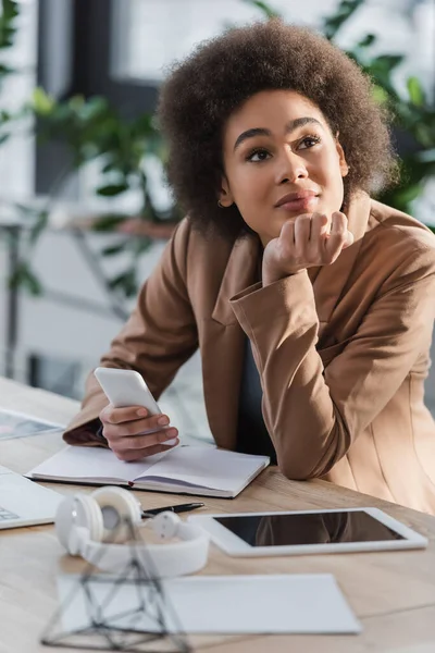 Dreamy mujer de negocios afroamericana sosteniendo teléfono inteligente cerca de dispositivos y auriculares en la oficina - foto de stock