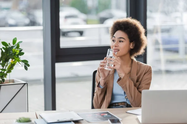 Sorridente donna d'affari afroamericana che tiene un bicchiere d'acqua vicino a dispositivi e carte in ufficio — Foto stock