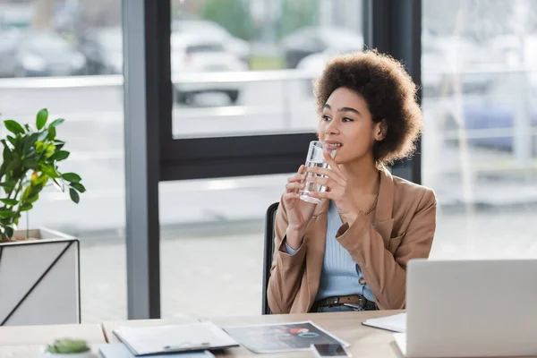 Femme d'affaires afro-américaine souriante tenant un verre d'eau près des appareils et des papiers au bureau — Photo de stock