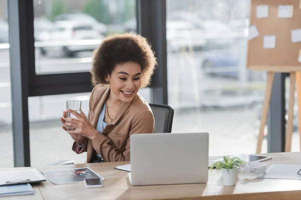 Enthousiaste femme d'affaires afro-américaine tenant un verre d'eau près d'un ordinateur portable et des documents au bureau — Photo de stock