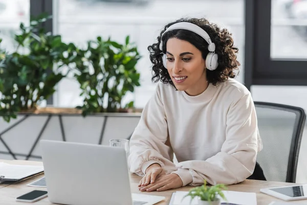 Mulher de negócios muçulmana sorridente em fones de ouvido olhando para laptop no escritório — Fotografia de Stock