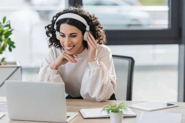 Cheerful muslim businesswoman in headphones looking at blurred laptop near notebook in office — стоковое фото