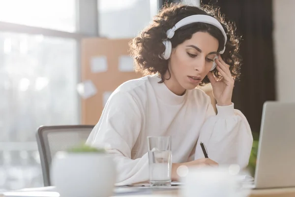 Arabische Geschäftsfrau mit Kopfhörer schreibt auf Notizbuch neben Laptop im Büro — Stockfoto