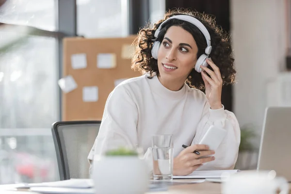 Fröhliche muslimische Geschäftsfrau mit Kopfhörer hält Stift und Handy im Büro — Stockfoto