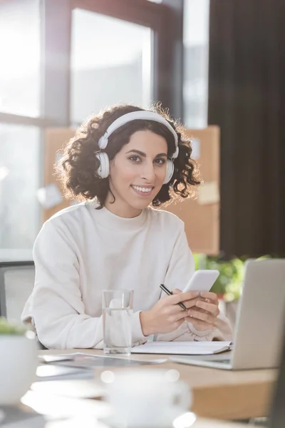 Усміхнена арабська бізнес-леді в навушниках тримає мобільний телефон і дивиться на камеру в офісі — стокове фото