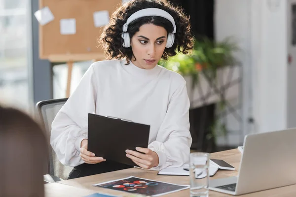 Empresaria musulmana en auriculares sujetando portapapeles cerca de la computadora portátil en la oficina - foto de stock