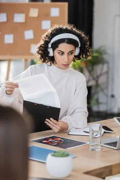 Mujer de negocios árabe en auriculares sujetando portapapeles cerca de dispositivos en la oficina - foto de stock