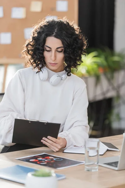 Young arabian businesswoman with headphones looking at clipboard near gadgets in office — Stockfoto