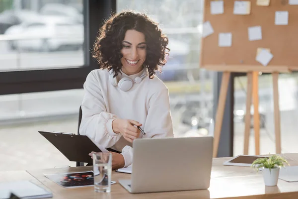 Cheerful muslim businesswoman with headphones holding clipboard during video call on laptop in office — Photo de stock