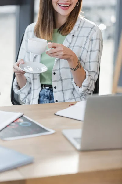 Ausgeschnittene Ansicht einer lächelnden Geschäftsfrau mit einer Tasse Kaffee in der Nähe eines verschwommenen Laptops im Büro — Stockfoto