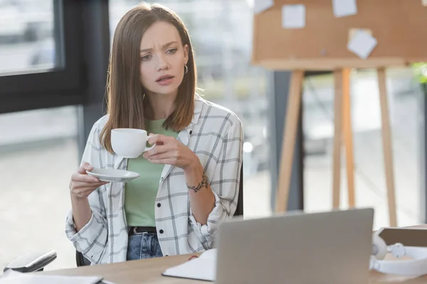 Орієнтована бізнес-леді тримає чашку біля ноутбука в офісі — стокове фото