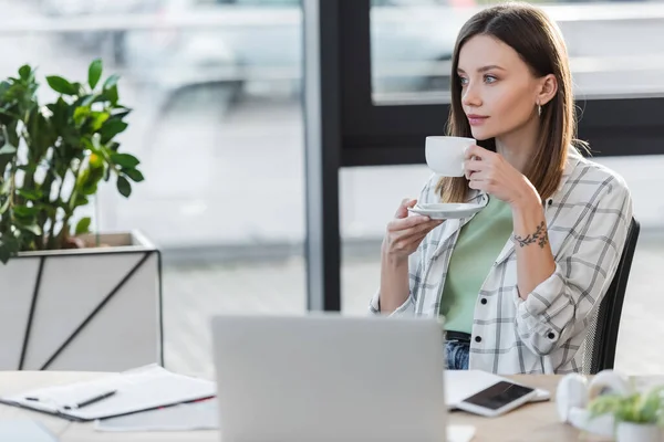 Jeune femme d'affaires tenant une tasse près des appareils flous dans le bureau — Photo de stock