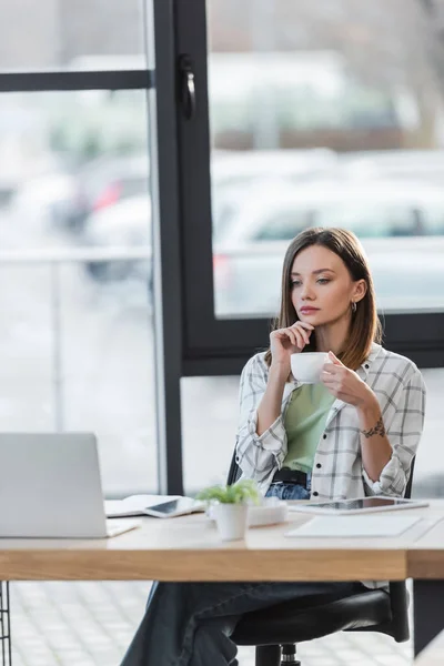 Femme d'affaires pensive tenant tasse près d'ordinateur portable flou dans le bureau — Photo de stock